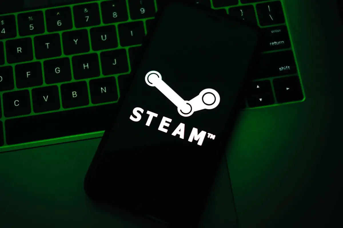 A smartphone screen prominently displaying the Steam logo, with a backlit laptop keyboard in the background emitting a green glow.