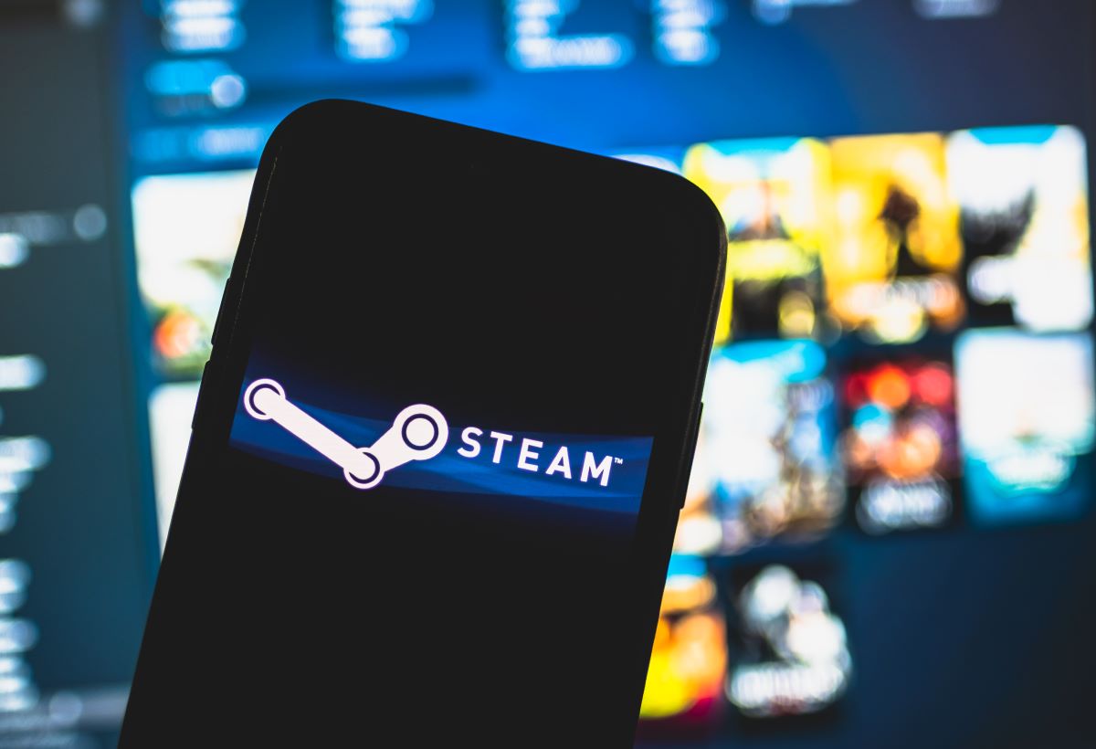 A smartphone displaying the Steam logo in focus, with a blurred background featuring what appears to be a monitor with various game graphics.