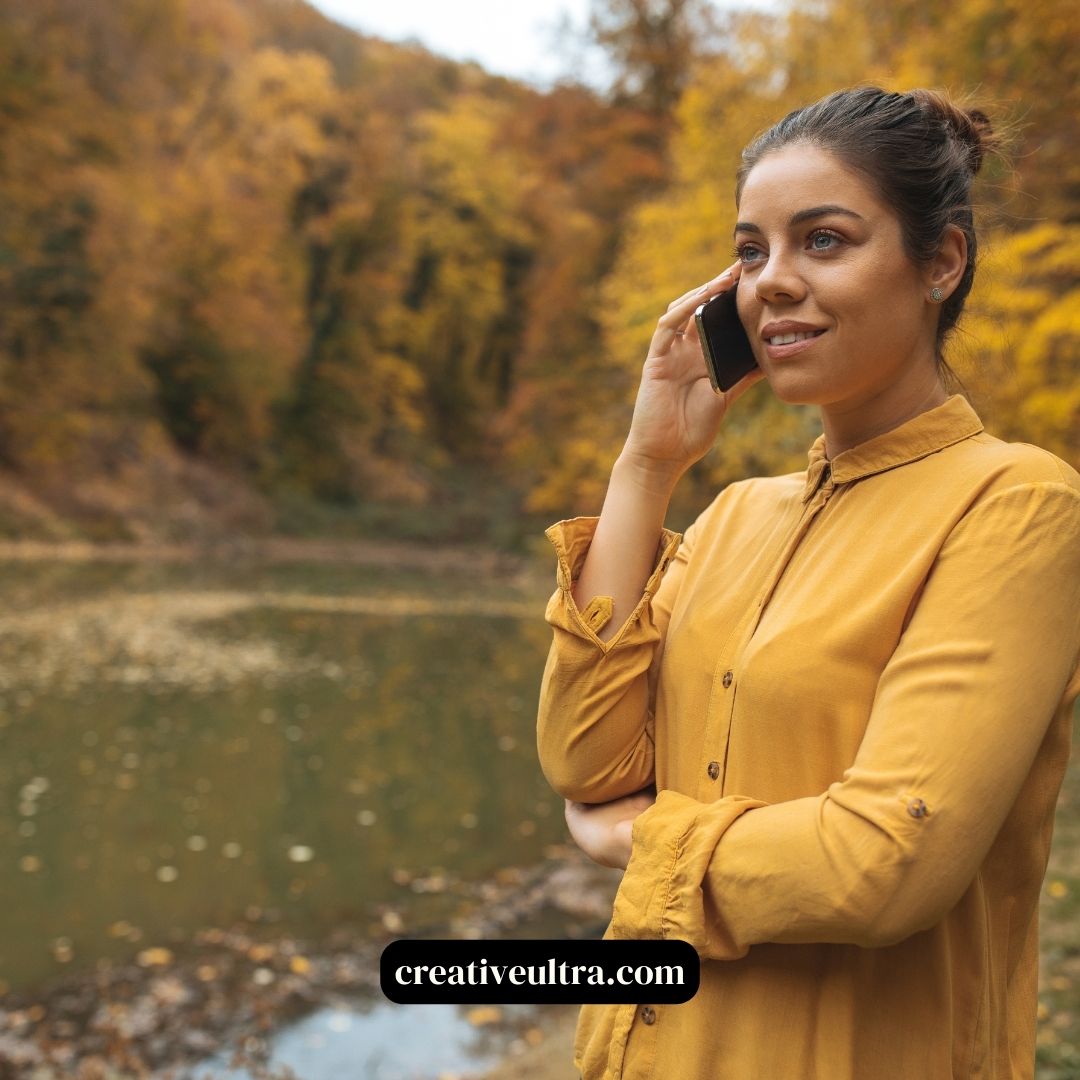 A woman in a mustard blouse smiles while talking on her phone, with a serene lake and vibrant autumn trees in the background, conveying a sense of calm.